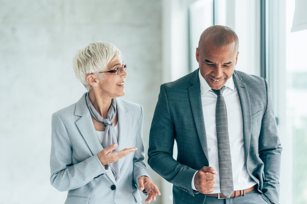 Two mature smiling business people walking through office hall and talking